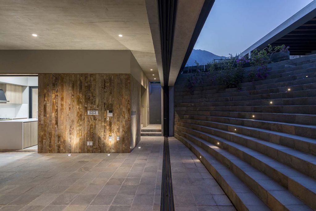 Dulanto realizó un meticulo trabajo de diseño de iluminación de la casa buscando realzar los materiales y creando una sofisticada atmósfera. Puertas corredizas de madera rústica permiten abrir la cocina al a´rea social. Se destaca el diseño con isla para mayor funcionalidad y aprovechamiento del espacio. Fotografía: Renzo Rebagliati. 